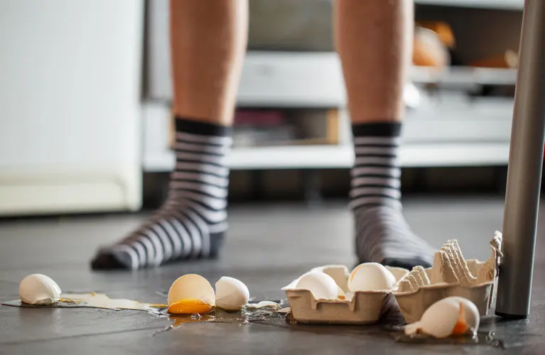 Cracked eggs spilled on a dark kitchen floor in front of a person wearing striped socks.