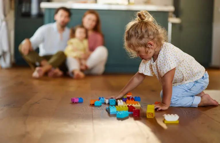 Little girl playing with building blocks on warm oak vinyl flooring with her family in the background.