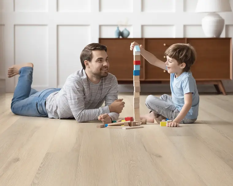 Father and son building a toy block tower on a light oak wood-look floor.