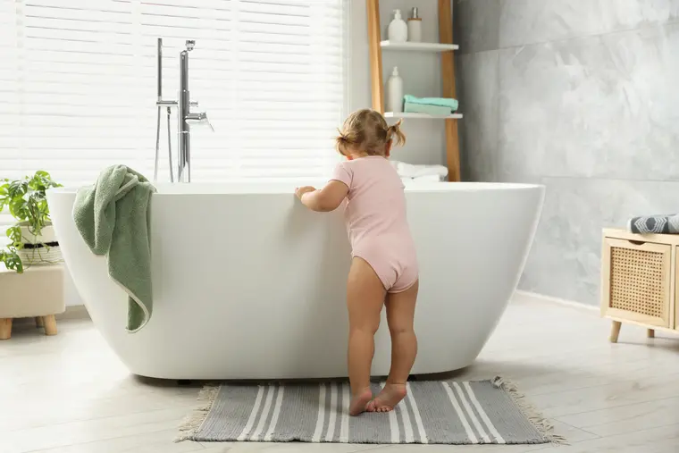 Toddler in a pink bodysuit standing next to a white freestanding bathtub on light tile floors.
